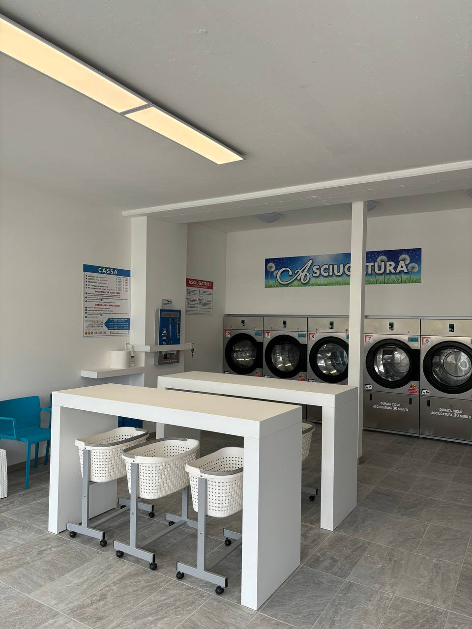 Interno della lavanderia con asciugatrici, tavoli e sedute